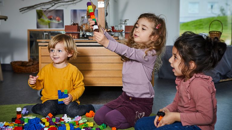 Tre barn sitter på golvet i en förskola och bygger med färgglada klossar. De ser glada ut.