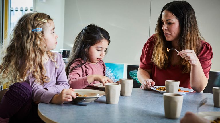 En pedagog och två barn sitter vid ett bord och äter. Det står koppar och fat på bordet.