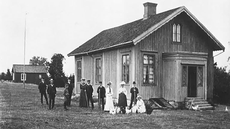 Historisk bild på Askimsskolan. Utanför den ursprungliga skolbyggnaden poserar en grupp välklädda män och kvinnor.
