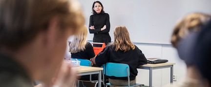 En lärare står vid tavlan och tittar ut över ett klassrum med elever.