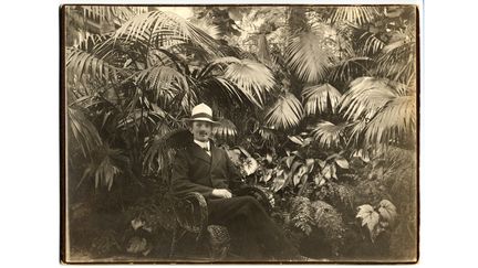 Äldre porträtt i sepiaton av en man som sitter i en korgstol med palmer i bakgrunden.