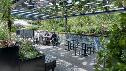 En grupp människor sitter runt ett bort på ett nedsänkt trädäck omgivet av grönska.