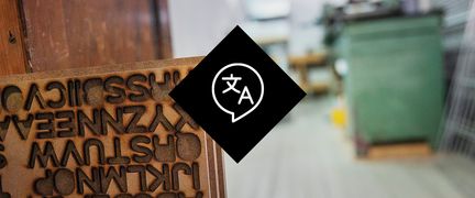 Fotografi, i förgrunden en skylt med bokstäver för tryck (typografi) och i bakgrunden en verkstad med arbetsbänk. I mitten en illustration med en pratbubbla med ett japanskt tecken och bokstaven A.