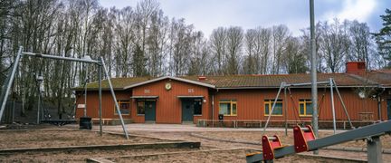 En röd skolbyggnad med en gungställning och en gungbräda framför.