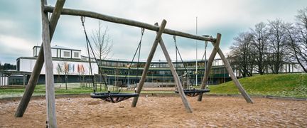 En gungställning av trä i en sandlåda på en skolgård med en skolbyggnad i bakgrunden.