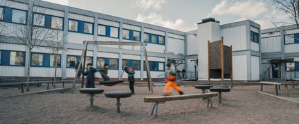 En grupp elever som leker på en skolgård med en sandlåda och en vit skolbyggnad i bakgrunden.