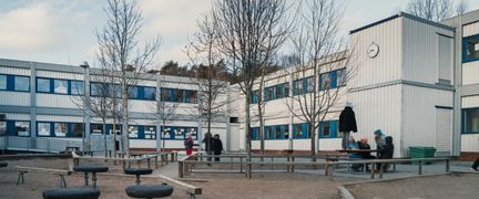 En grupp elever sitter på bänkar och står framför en vit skolbyggnad.