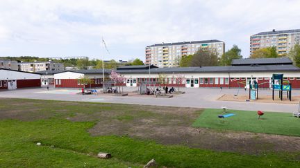 En skolgård med grön gräsmatta och lekställningar framför den röda skolbyggnaden. Bakom skolbyggnaden syns hustak från ett bostadsområde.
