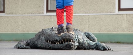 En bronsskulptur av en krokodil ligger på marken. Ovanpå syns benen på ett barn som har klättrat upp på krokodilen.