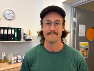Porträttbild på Niklas. Han har runda glasögon, mustasch, grön tröja och keps. 