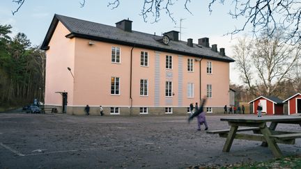 Huvudbyggnaden för Högenskolan med en parkbänk och några barn i bakgrunden.
