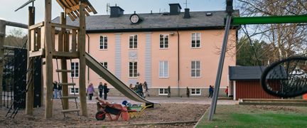 En klätterställning med en rutschkana på en lekplats framför Högenskolan. En gunga och några barn syns även i bakgrunden.