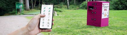 En hand som håller i en mobiltelefon med Piffl-appen igång. En rosa box i bakgrunden.