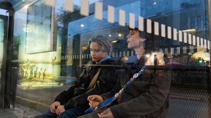 Två ungdomar sitter i en busskur, vi ser dem genom glaset.