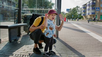 Ett barn och en vuxen som vinkar hejdå vid en busshållsplats.