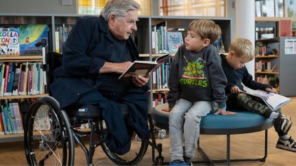 En person utan ben sitter i rullstol i ett bibliotek och läser för ett barn.