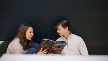 Två personer sitter mot en svart vägg och tittar i en bok.