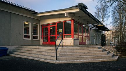 En grå skolbyggnad med röd entré med trappor som leder upp till den.