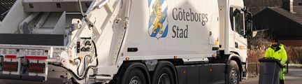 En vit sopbil med Göteborgs Stads logotyp på sidan. En sophämtare i gula arbetslkäder går med ett grönt kärl vid sopbilen.