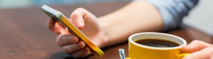 En person håller en mobiltelfon i handen till vänster och en kaffekopp i handen till höger. 