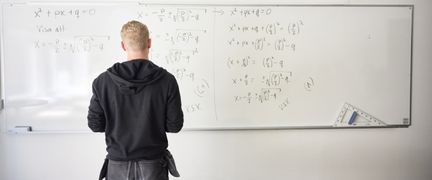 En kille i snickarkläder står framför en tavla med mattetal.