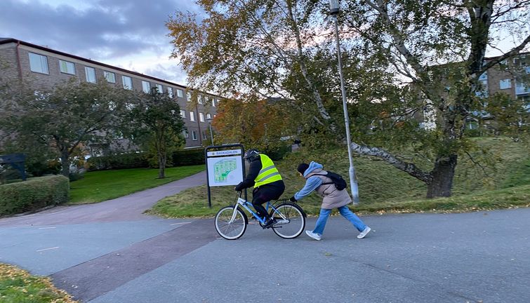 En person med gul väst på cykel och en annan som puttar cykeln bakifrån