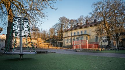 En karusell, gungor och en lekstuga på en skolgård framför ett skolhus i trä