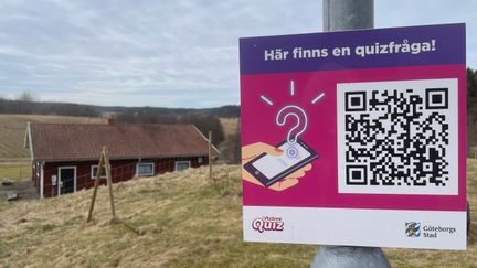 En skylt med texten Här finns en quizfråga och en QR-kod. I bakgrunden syns fält och ett rött hus.