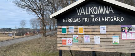 Skylt med texten Välkomna till Bergums fritidslantgård. I bakgrunden syns träd, en väg och röda hus.