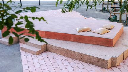 Göteborgs hbtqi-monument skymtar bakom ett träd. Verket består av rosa marksten och två lager i rosa betong, med tre kuddar i marmor som ligger på det översta lagret.