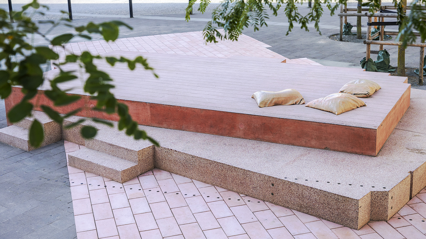 I förgrunden syns löv på träd, bakom ser man monumentet: tre rosa lager i marksten och betong. Överst ligger tre kuddar i marmor.
