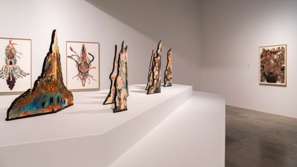 Installationsbild med färgglada spetsiga skulpturer i förgrunden och tre stora akvareller i bakgrunden.