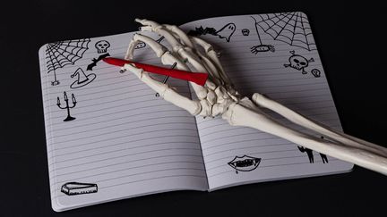 En skeletthand håller i en röd penna över en uppslagen anteckningsbok med teckningar av bland annat spindelnät, dödskallar och fladdermöss.