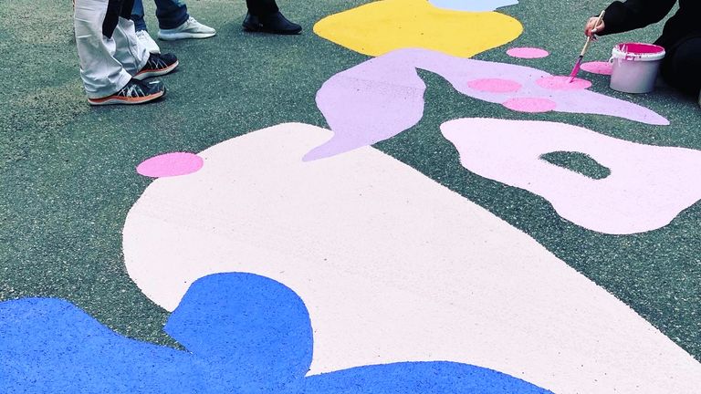 Bild på målningar på asfalten på Gröna Gatan. Färgglada blommor och prickar syns på denna bild. Det syns även en färgbunke och en mnniskas hand med en pensel. Bredvid syns fötterna på tre personer som står bredvid den som målar. 