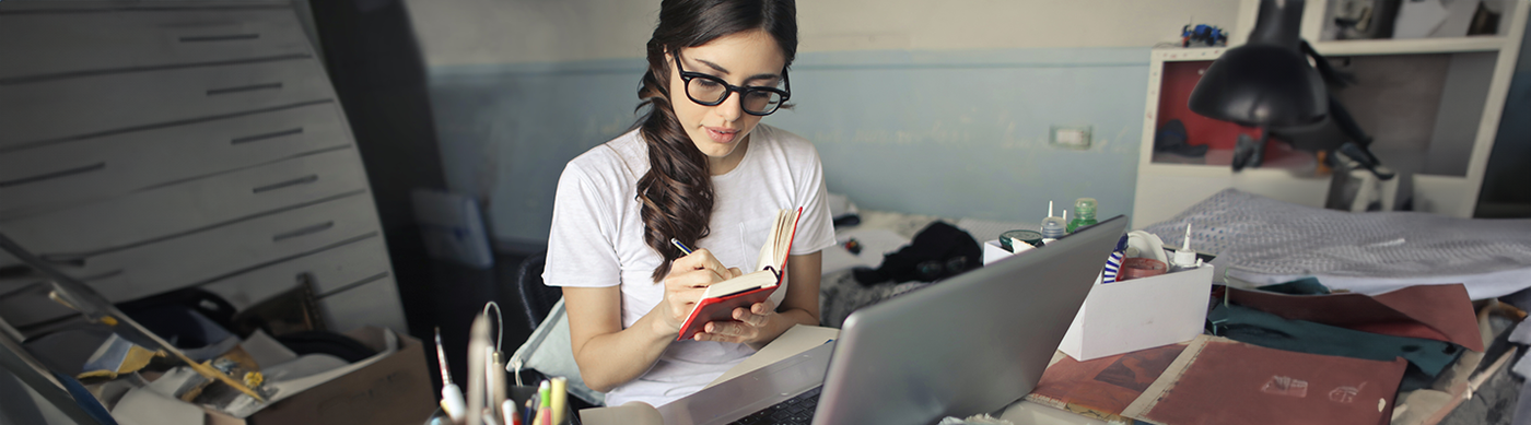 En kvinna sitter vid ett skrivbord och för anteckningar i en anteckningsbok. På skrivbordet står det en dator.