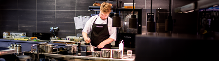 En man i kockkläder står i ett restaurangkök och rör i en gryta.