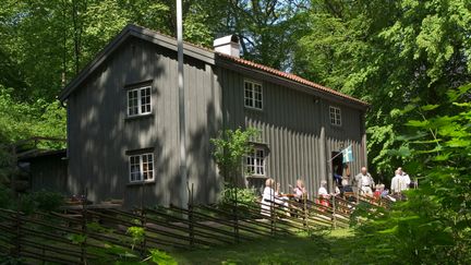 Ett stort grått trähus med människor som sitter på bänkar framför.