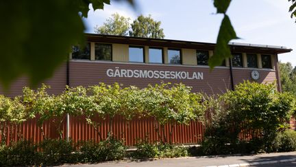 En röd skolbyggnad med texten Gärdsmosseskolan på fasaden. Gröna buskar och löv syns i bild.