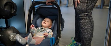 Bebis i babyskydd vid sidan av vikter på gymmet. En vuxen persons ben syns bredvid.