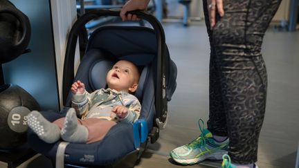 Bebis i babyskydd vid sidan av vikter på gymmet. En vuxen persons ben syns bredvid.