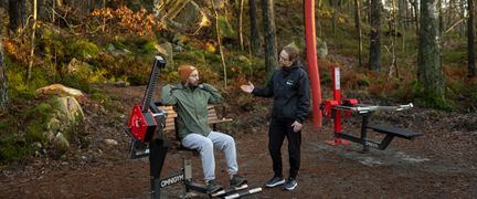 En person i en träningsmaskin i ett utegym. En instruktör står vid sidan och pratar med personen som tränar.