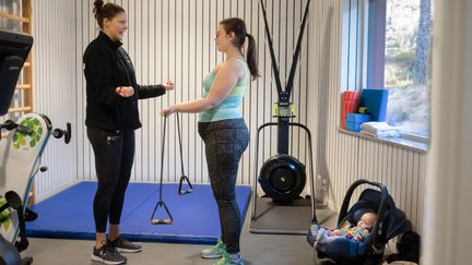 Två personer i gymmet med en bebis i babyskydd bakom dem.