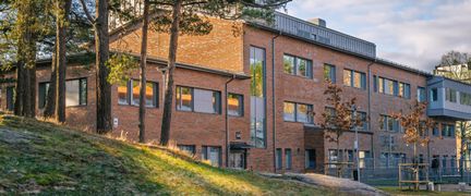 En röd skolbyggnad i tegel som ligger bakom en kulle med träd.