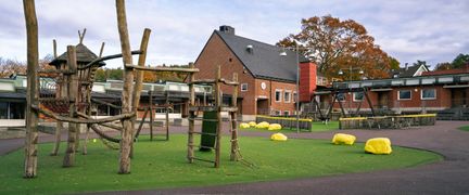 En lekplats med ett par gungor och en klätterställning framför Hovåsskolan. I bakgrunden syns ett par gulmålade stenar.