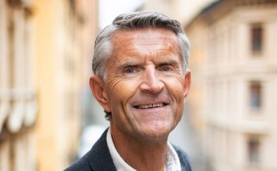 Ansikte på man som ler. Han har kortklippt grått hår. Gula husfasader syns i den oskarpa bakgrunden.