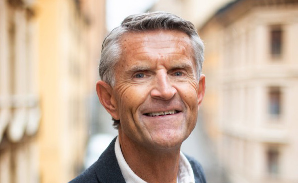 Ansikte på man som ler. Han har kortklippt grått hår. Gula husfasader syns i den oskarpa bakgrunden.