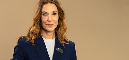Direktören står mot en beige bakgrundsbild. Hon tittar in i kameran. Hon har på sig en blå kavaj med en vit tröja under. 