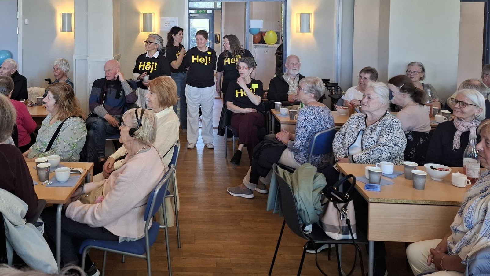 Många deltagare i ett stort rum tittar antagligen på en föreläsare. I bakgrunden står några funktionärer med "hej" på tröjan.