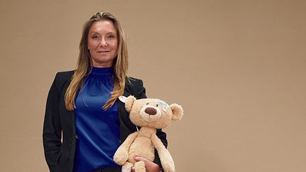 En person står mot en beige bakgrund. Hål tittar in i kameran och har ena handen i byxfickan. I den andra handen håller hon i en nallebjörn.