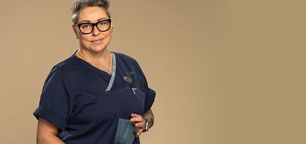 En person står mot en beige bakgrundsvägg. Hon har på sig mörkblåa vårdkläder. Hon tittar in i kameran och har svarta glasögon på sig.